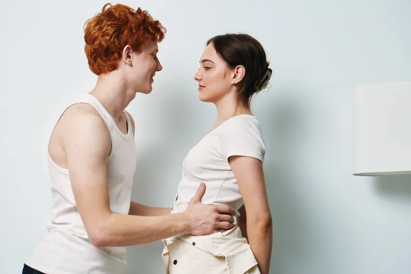 Young couple, man and woman standing close, looking at each other with soft expressions in casual clothes indoors. Intimate moment, connection and gentle interaction between two people.