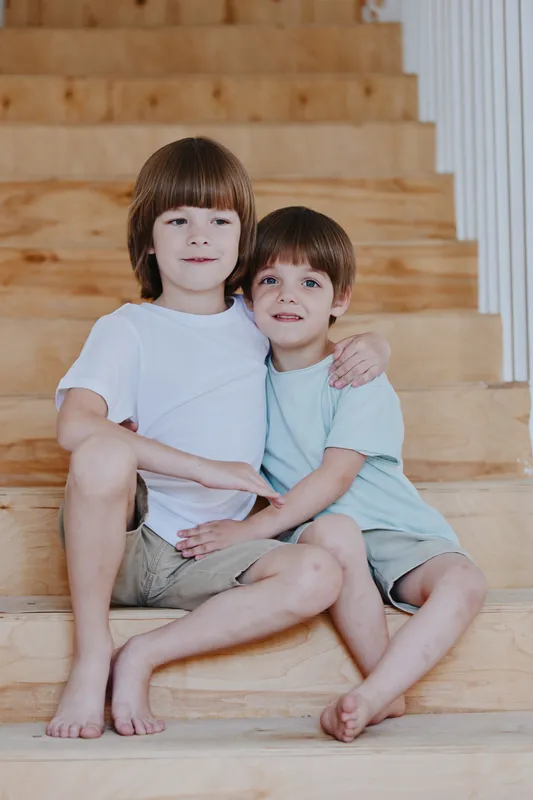 Two happy boys sitting on wooden stairs, embracing fondly, showcasing warmth and brotherly love in a bright, cheerful setting with natural light.