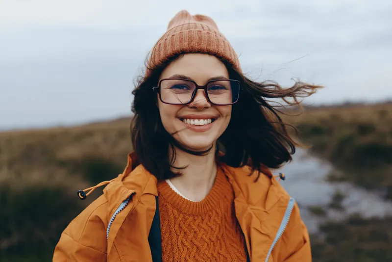 Happy woman lifestyle outdoors, smiling in warm hat and glasses, wearing jacket and knit sweater, hair moved by wind on a coastal path, soft natural light, relaxed travel mood