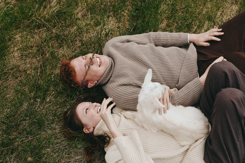 Couple outdoors, lying on grass, cozy sweaters, affectionate moment, goat kid nearby, warm sunlight, relaxed lifestyle, candid and natural colors, peaceful scene, close companionship, light breeze,