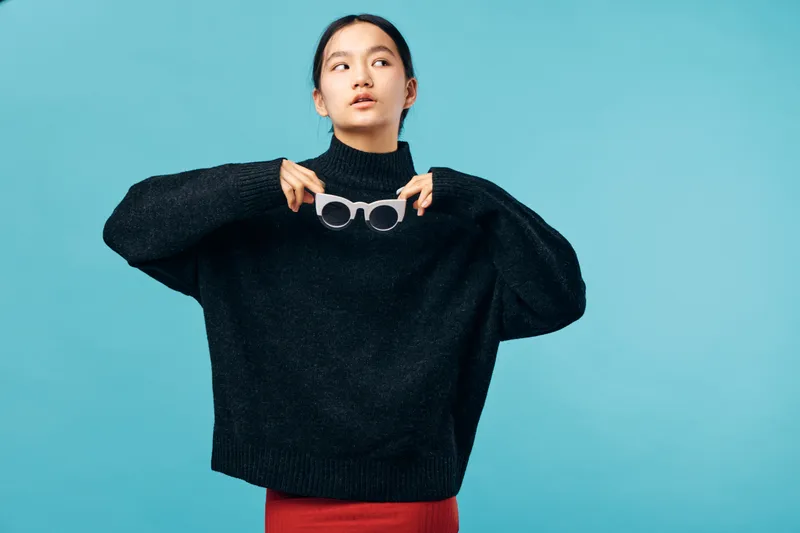 Young woman wearing black sweater holding sunglasses with both hands looking sideways on blue background. Casual fashion style with thoughtful expression and minimalist studio setup.