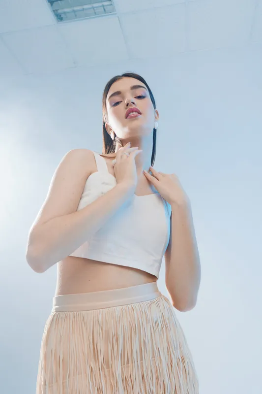 Young woman in white top and beige skirt poses confidently against a light background. Concept of beauty, fashion, and modern style with soft natural lighting.