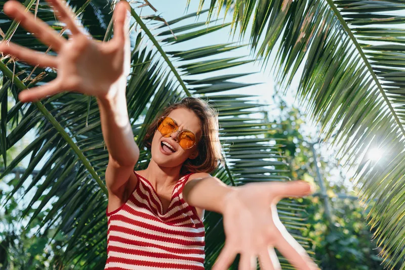 A cheerful woman wearing a striped sleeveless top smiles broadly and reaches toward the camera in a sunlit tropical setting, exuding energy, joy, and a carefree summer vibe.