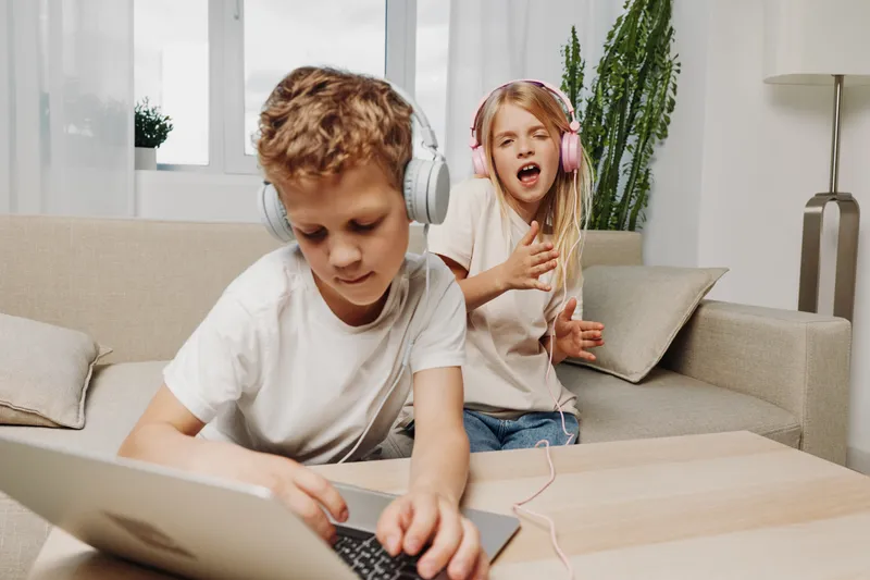Two children with headphones enjoying music at home, girl singing passionately and boy focused on tablet. Bright modern living room setting with casual clothing and natural light.