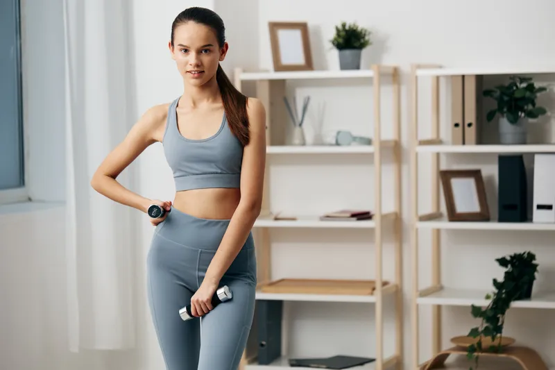 Fitness woman in athletic wear stands with dumbbells at a home gym, performing a straightforward workout. Bright room with shelves and plants, highlighting training, health and personal progress.