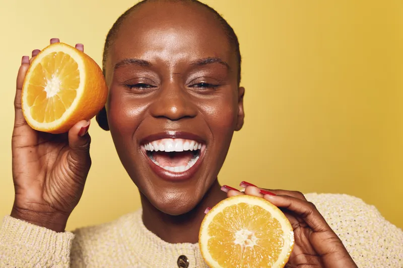 Smiling black woman with dark skin holds two halves of fresh orange fruit near face. She wears beige sweater and poses on yellow background. Concept of healthy lifestyle and natural beauty.