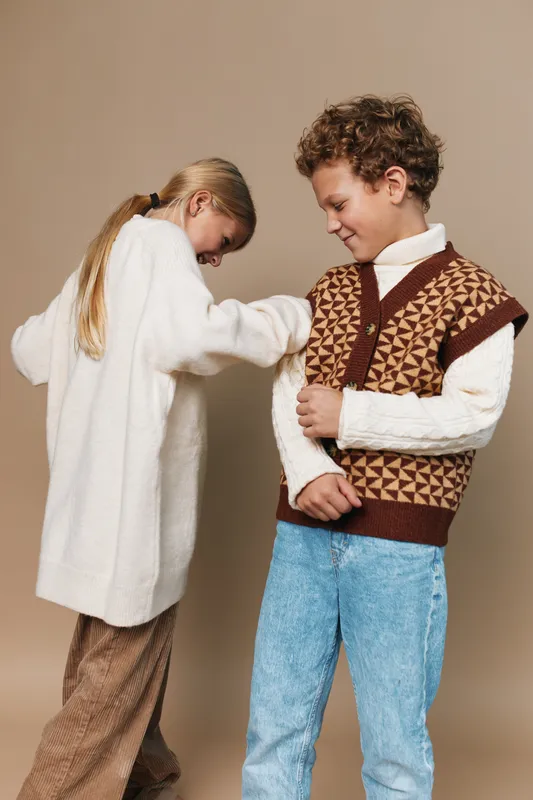 Two children share a playful moment in vintage style outfits, as a girl helps a boy adjust his knitted vest, creating a warm, wholesome scene full of friendship and charm.