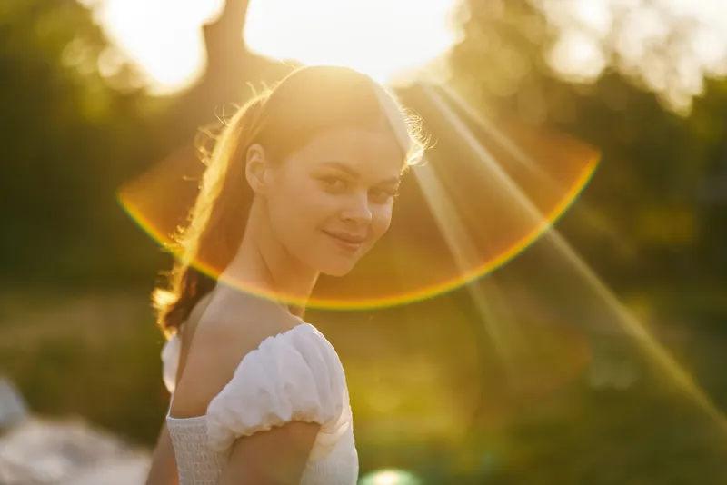 Young woman with a serene expression enjoying a sunny day in nature, wearing a white dress, surrounded by a soft golden glow and vibrant greenery