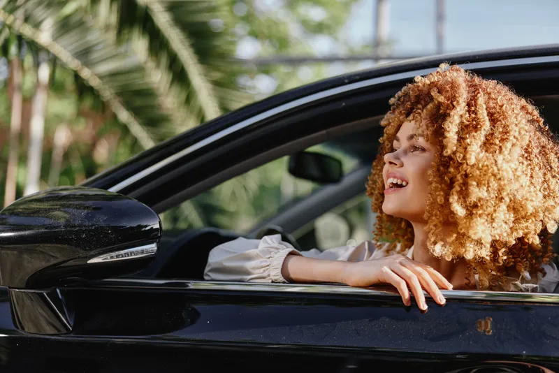 smiling woman with curly hair leaning out of a black car window, enjoying sunny weather and tropical palm trees in the background