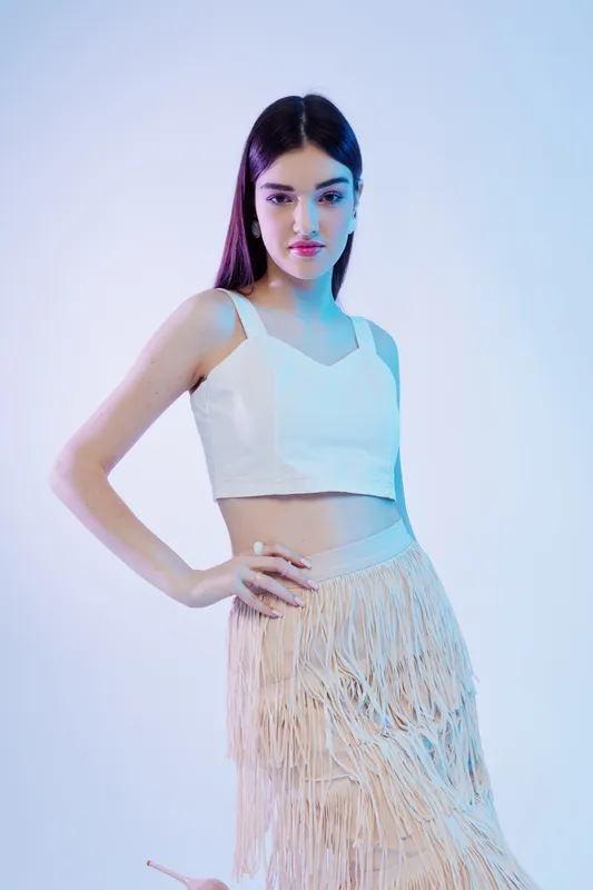 Young woman in white top and fringed skirt poses confidently against a light background. Concept of woman beauty fashion with modern style and elegance.