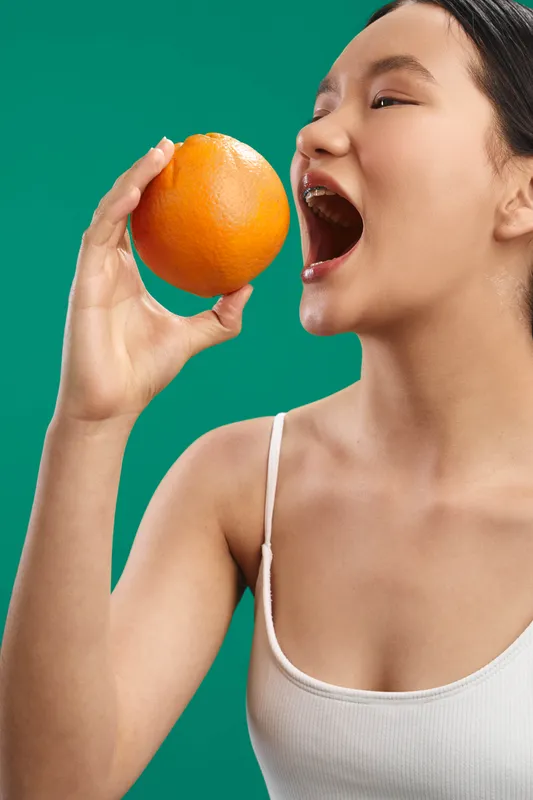 Asian woman enjoys a bright moment as she bites into a ripe orange, conveying playful energy and casual refreshment in a vibrant, contemporary setting.