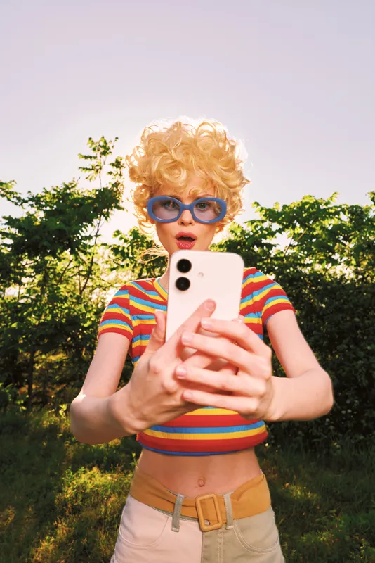 Cheerful woman outdoors, taking a selfie, retro style, colorful striped top, oversized sunglasses, curly blonde hair, sunlit garden backdrop, casual lifestyle moment, natural colors