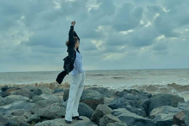 Empowerment and victory on a rocky shore as a woman raises a fist toward the wind, facing the sea, symbolizing determination, resilience and freedom in a bold outdoor scene