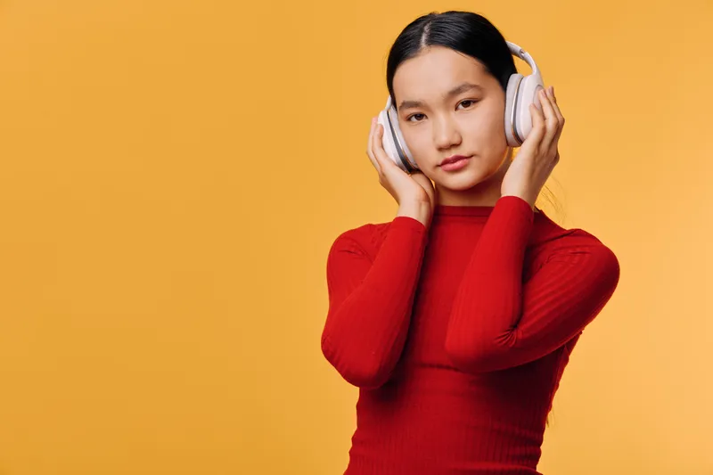 Young woman wearing headphones and red sweater listens to music with calm expression against orange background in studio. Asian girl with modern casual style enjoying audio technology.