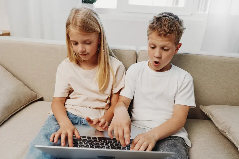 Curious children sitting on a couch using a laptop together, exploring digital content. Bright room with natural light creates a cozy atmosphere for learning and entertainment.