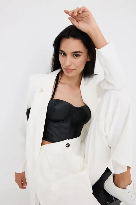 Young woman in white suit and black corset with confident expression posing against plain white background in modern fashion style with natural makeup and dark hair.