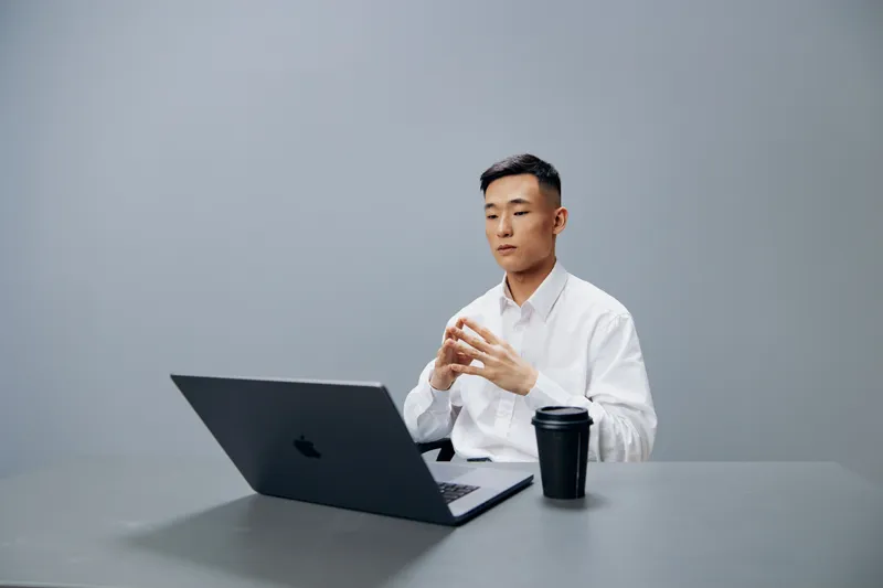Мужчина азиатской внешности испытывает усталость и эмоции, работая за ноутбуком с чашкой кофе на рабочем столе руководителя