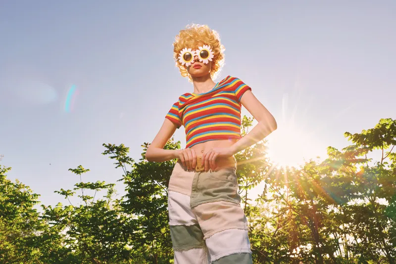 A stylish woman poses outdoors in a sunny park, wearing a striped tee and round floral sunglasses. Retro film color tones evoke carefree summer vibes and playful fashion.