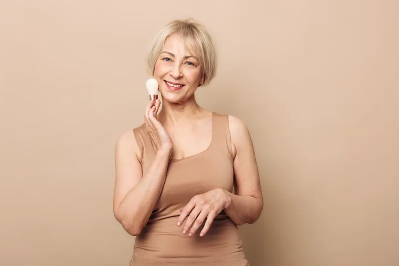 Smiling mature woman with short blonde hair uses facial cleansing brush on beige background. Concept of skincare, beauty routine, anti aging, and self care for older women.