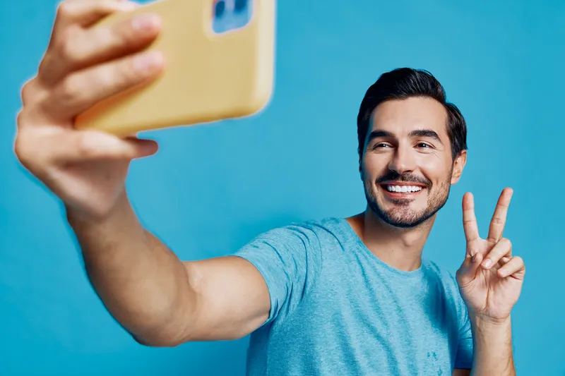Happy young man taking a selfie with smartphone, smiling brightly, wearing casual blue t-shirt, posed against a vibrant blue background. Fun lifestyle concept.