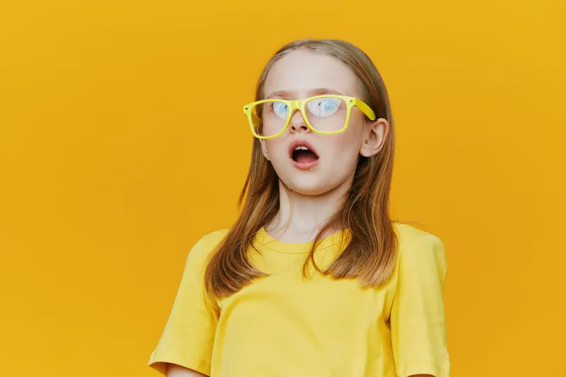 A surprised young girl with glasses poses against a vibrant yellow background, wearing a bright yellow t shirt, embodying joy and curiosity in a playful setting