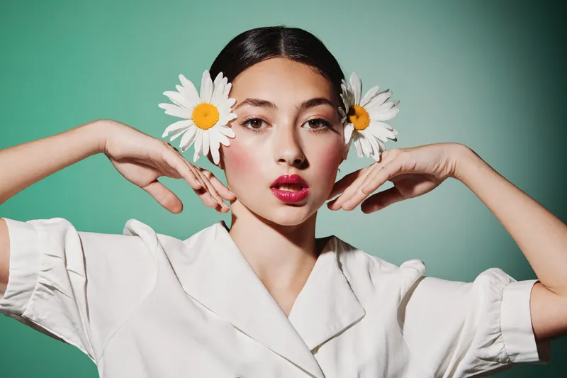 Fashionable young woman with daisy flowers in her hair poses against green background. Bright makeup and white outfit create artistic beauty concept and fresh creative style.
