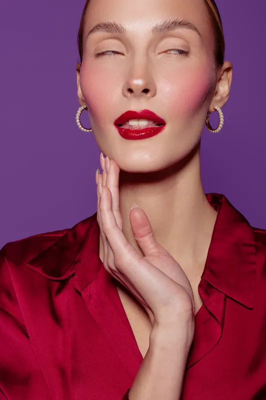 Close-up portrait of a woman with red lipstick and hoop earrings wearing a red blouse on purple background. Concept of beauty, fashion, makeup, and elegance in studio lighting.