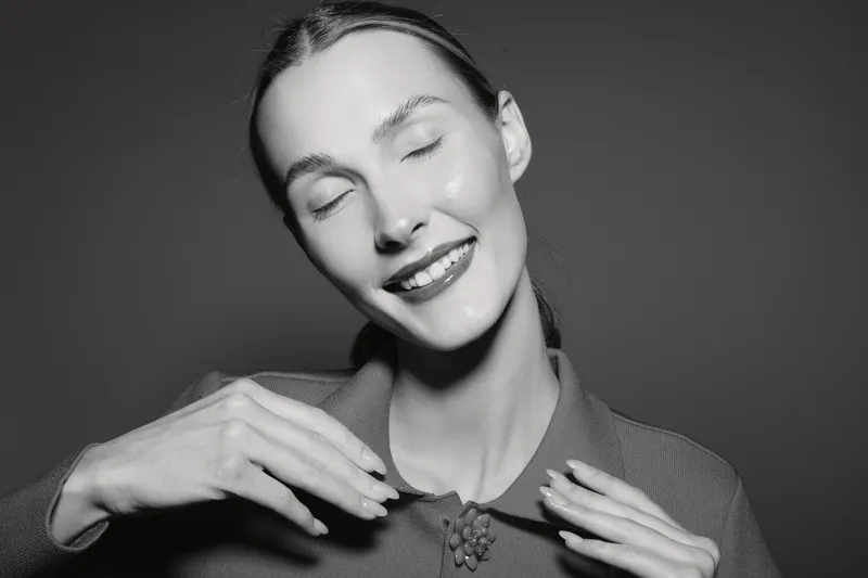 Smiling white woman with light skin and closed eyes enjoys moment with hands near neck in monochrome studio portrait.