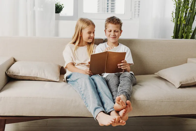 Two happy children sitting barefoot on a beige sofa reading a brown book together in a bright modern living room. Concept of childhood, learning, family bonding, and home comfort.