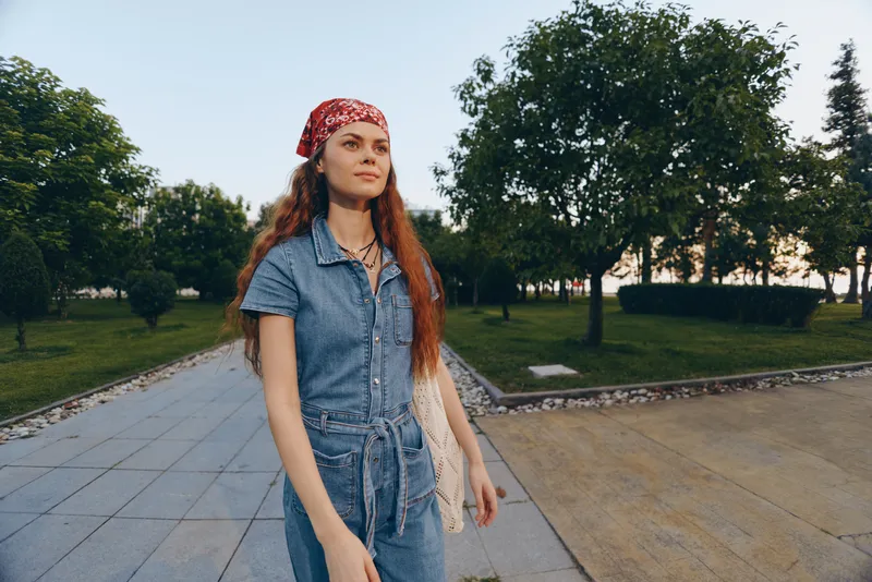 Lifestyle woman in modern boho-western denim street style wearing a red bandana outdoors with film color tones and natural green park background.