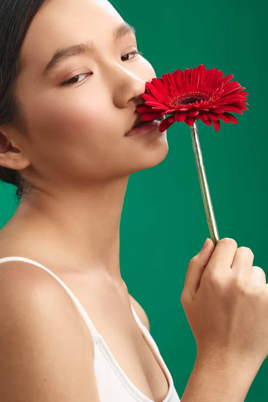 Asian woman gracefully holds a red flower close to her lips, conveying a serene mood amid a vivid green backdrop. The composition blends beauty, elegance, and natural poise in a refined studio