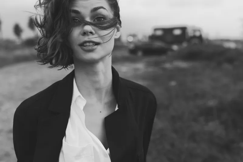 Female in blazer and white shirt outdoors, wind blowing hair, moody black and white aesthetic, professional yet casual look, open field setting with distant vehicle, dramatic atmosphere and modern