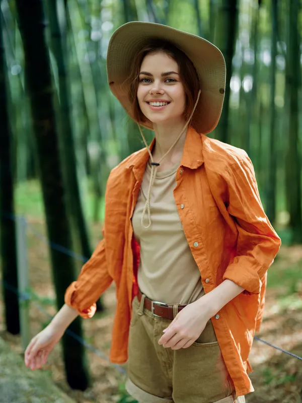 Woman smiling in a straw hat walking in a bamboo forest, nature portrait outdoors with casual orange shirt and shorts, travel style, relaxed mood and sunlit green background.