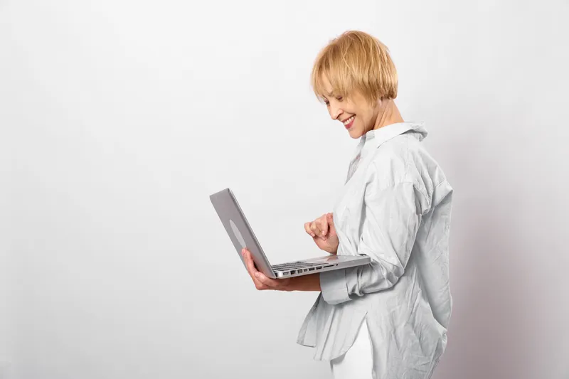 Woman with laptop, smiling in studio, white coat, focused on work, using technology for communication, relaxed posture, short blonde hair, clean background