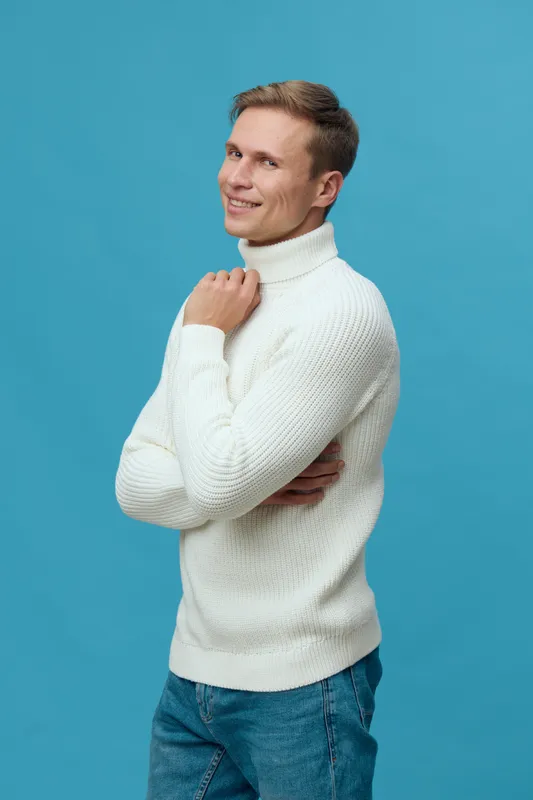 Smiling young man with light brown hair wearing a cozy white turtleneck sweater posing against a solid blue background. Casual style, happy expression, photo studio portrait, lifestyle concept, warm c