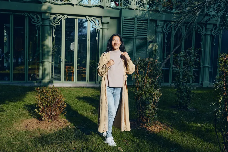 Woman outdoor coat fashion casual garden gate sunlight greenery breeze, standing on lawn wearing beige long coat, denim jeans and white sneakers, relaxed pose against a decorative fence in soft