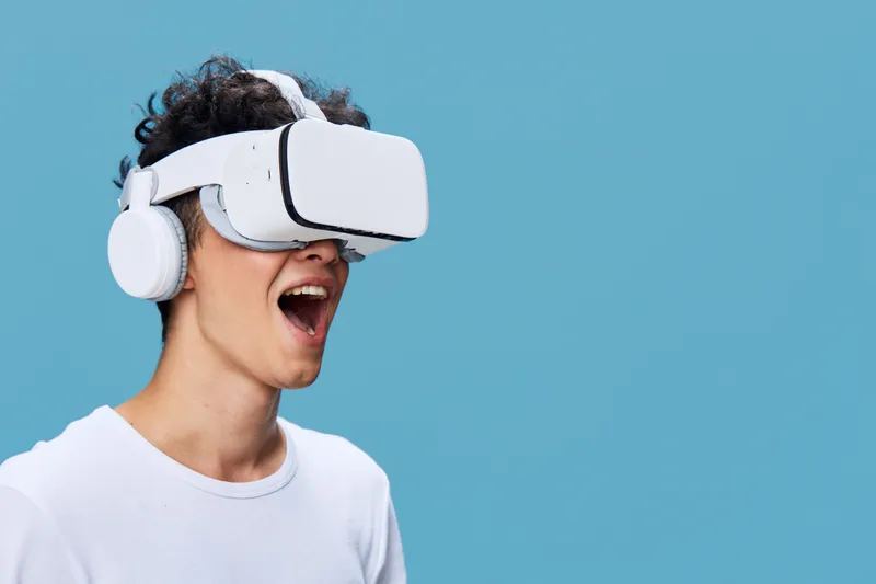 young man wearing virtual reality headset and headphones enjoying immersive digital experience against blue background with casual white t-shirt
