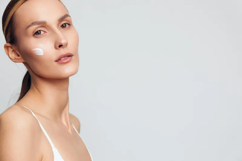 A young woman with clear skin applies white cream on her cheek while looking at the camera on a plain light gray background. Concept for skincare routine and natural beauty.