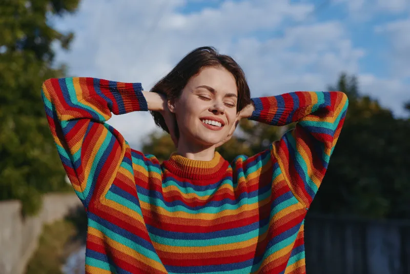 Woman young woman in a colorful striped sweater smiling with eyes closed outdoors, casual outfit and relaxed pose under warm sunlight on a suburban path, lifestyle fashion portrait.