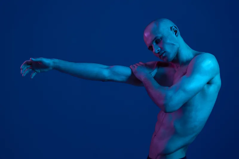 Confident man stretching his arm in a blue light setting, showcasing muscular features and a contemplative expression, perfect for fitness and wellness themes.