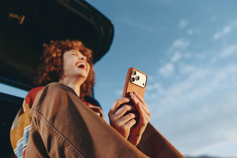 Woman smartphone lifestyle outdoors, curly haired woman in brown jacket smiles while holding a smartphone near a car, looking up at blue sky, candid travel moment during sunny day solo trip
