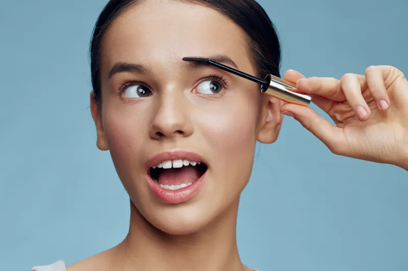 Cheerful young woman applying eyebrow gel against a light blue background, showcasing natural beauty and makeup techniques for a fresh look