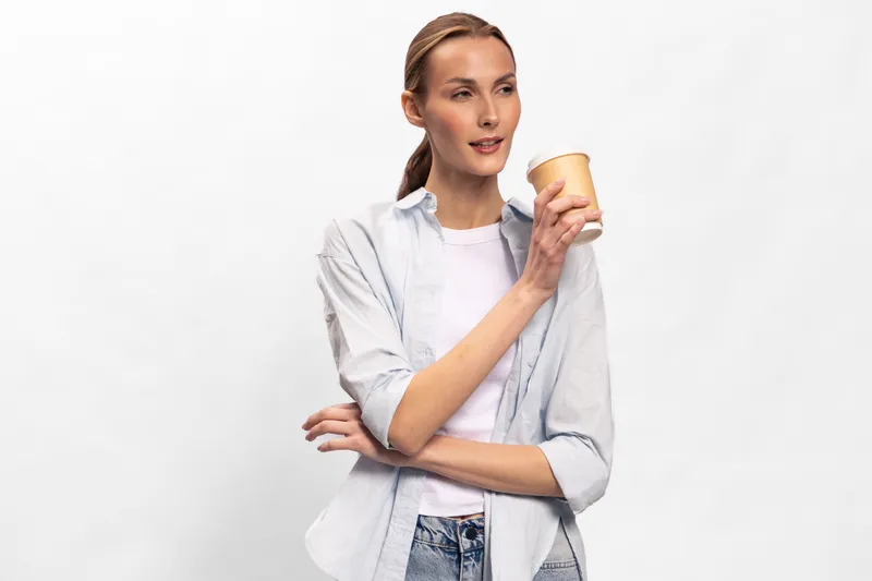 Young woman with ponytail holding disposable coffee cup looks away thoughtfully on white background. Casual clothing, relaxed mood, concept of coffee break and everyday lifestyle.