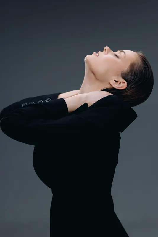Elegant woman in black outfit with closed eyes and serene expression, posing gracefully against gray background.