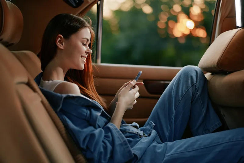 Young woman enjoying a relaxed moment in a car, wearing denim clothing, smiling while using her smartphone, with a soft sunset background.