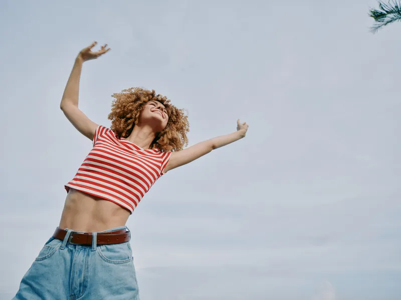 Young woman with curly hair joyfully raises arms in the air, wearing a red striped crop top and blue jeans, captured against a bright daytime sky.