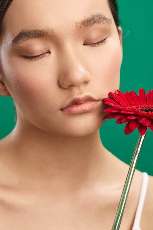 asian woman with a vibrant red flower near her face, calm expression, soft studio lighting creates a tranquil, elegant portrait that emphasizes beauty and grace.