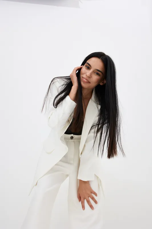 Smiling woman in white suit poses confidently with long dark hair and relaxed expression against a clean white background, showcasing modern fashion style.