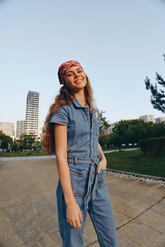 Lifestyle woman in modern boho-western denim street style wearing bandana, smiling confidently outdoors under clear sky with urban background, film color tone.