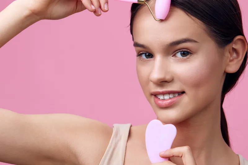 Young woman using a facial roller on her forehead, smiling, promoting self care and beauty routines, in a soft pink background with a modern aesthetic